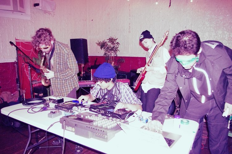 A sepia tinted photograph of 4 people in a large white and red room. From left to right: a curly-haired person in a flannel shirt, slightly unbuttoned is slouched over playing a saxophone; another person in a beanie and flannel is seated at a large white folding table twisting knobs on various electronic music devices; next a person in a white dress shirt, beanie, and cargo vest plays a large red bass guitar; lastly a person in an all black tracksuit and black coat with green tape for glasses is crouched over a laptop on the same white table. 