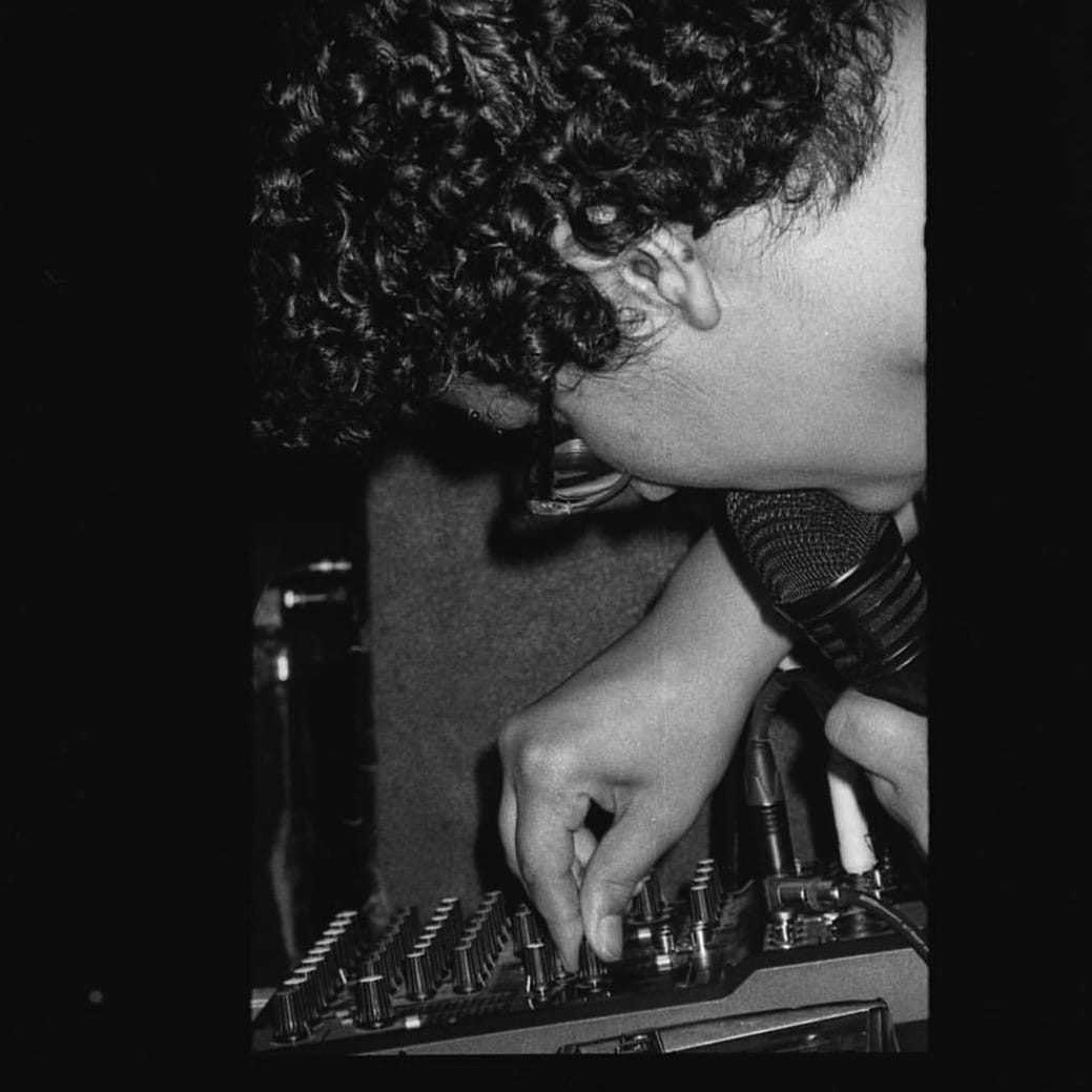 a close-up profile shot of a person with short curly hair and oval glasses holding up a microphone to their lips and twisting a knob on a audio mixer. The image is in black and white.