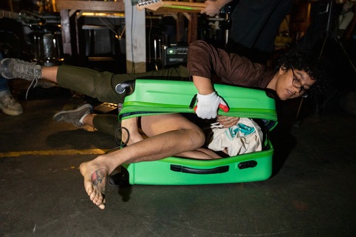 A landscape portrait of a person balancing atop a green suitcase, pulling it open with a gloved hand to expose the legs and hand of a person inside the suitcase. In the backgroun we can see a hand holding the neck of a guitar.