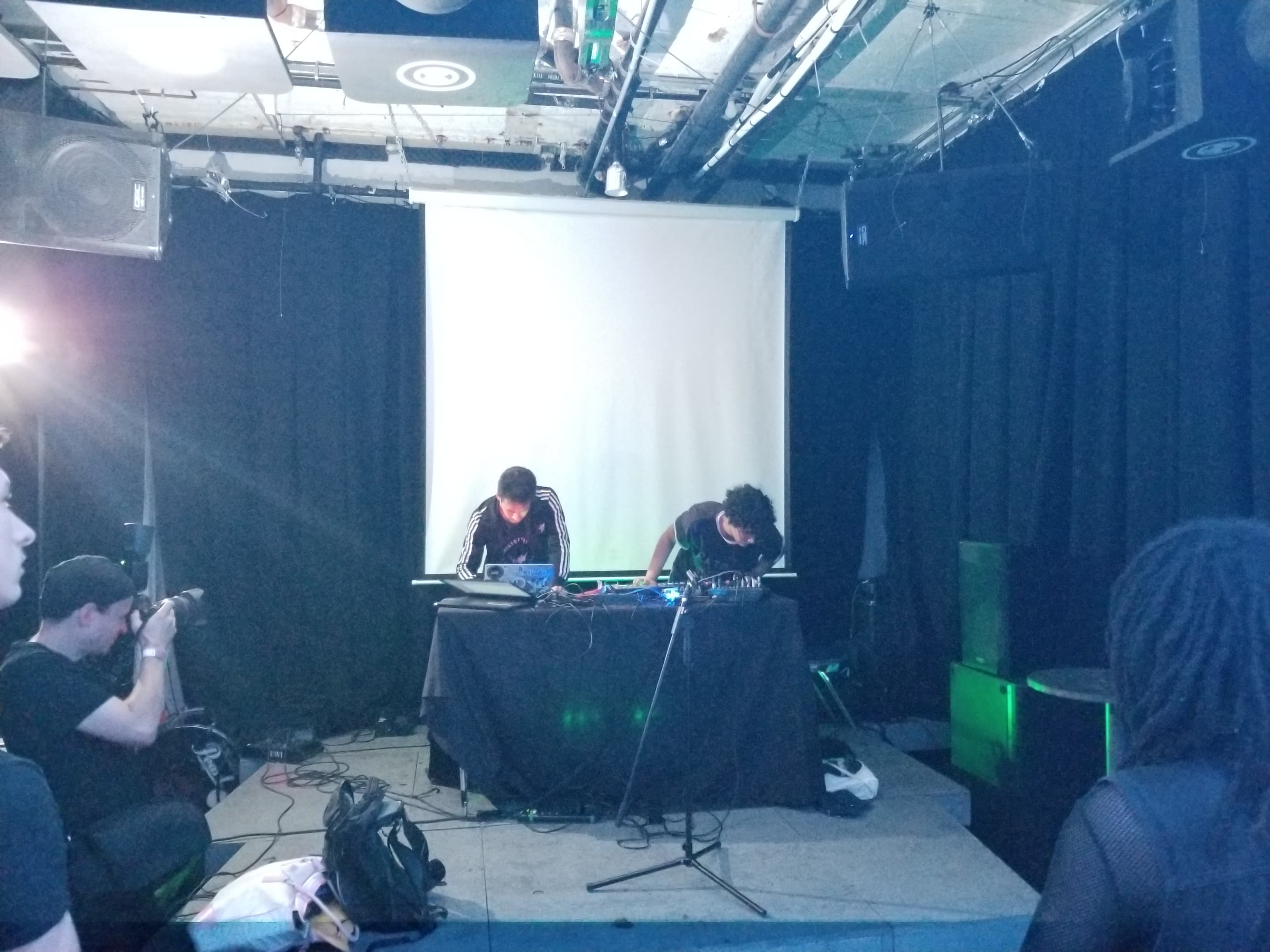 A photo of two people wearing black and white clotching at a table draped in black cloth on a stage in front of a small crowd in a medium sized performance room. On the table is a laptop and audio mixer as well as other devices and cables.