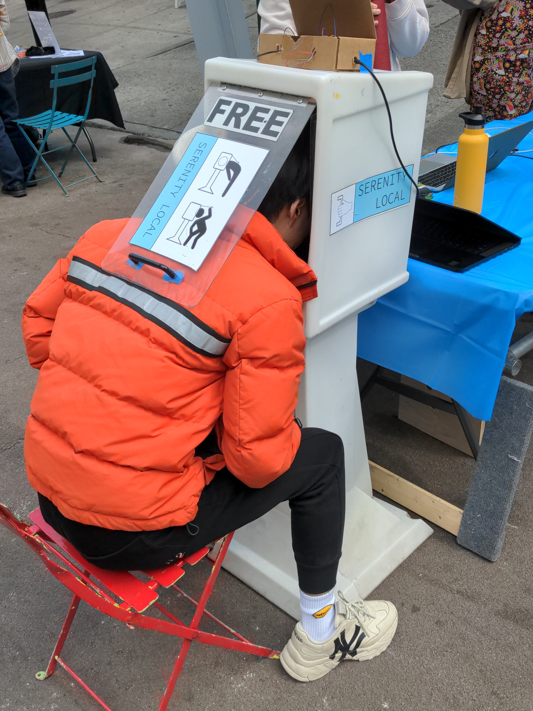 Image of a person in an orange puffer coat seated before a newspaper box in which they've stuck their head into. They are outdoors and there is daylight outside. They wear white sneakers with the logo of the New York Yankees on the side.
