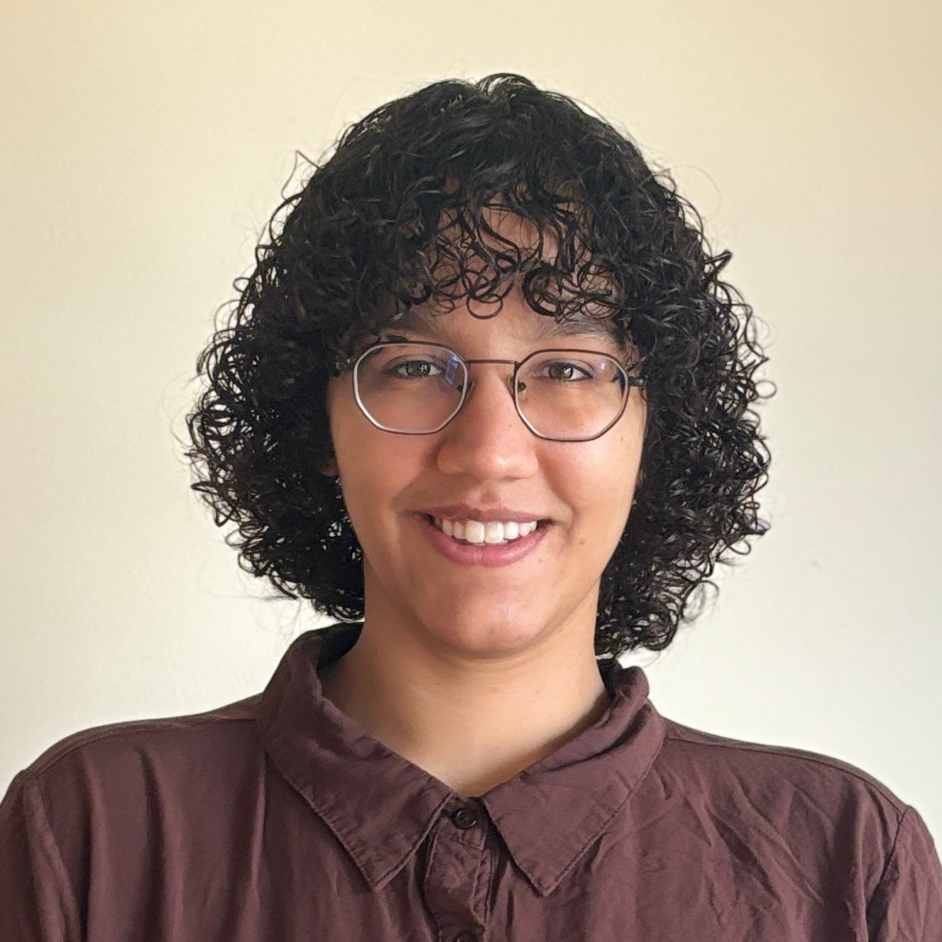 Portrait image of a lightskinned brown person with chin legnth curly hair
    and geometric black metal eyeglasses wearing a slighlty wrinkled brown button up shirt while smiling.